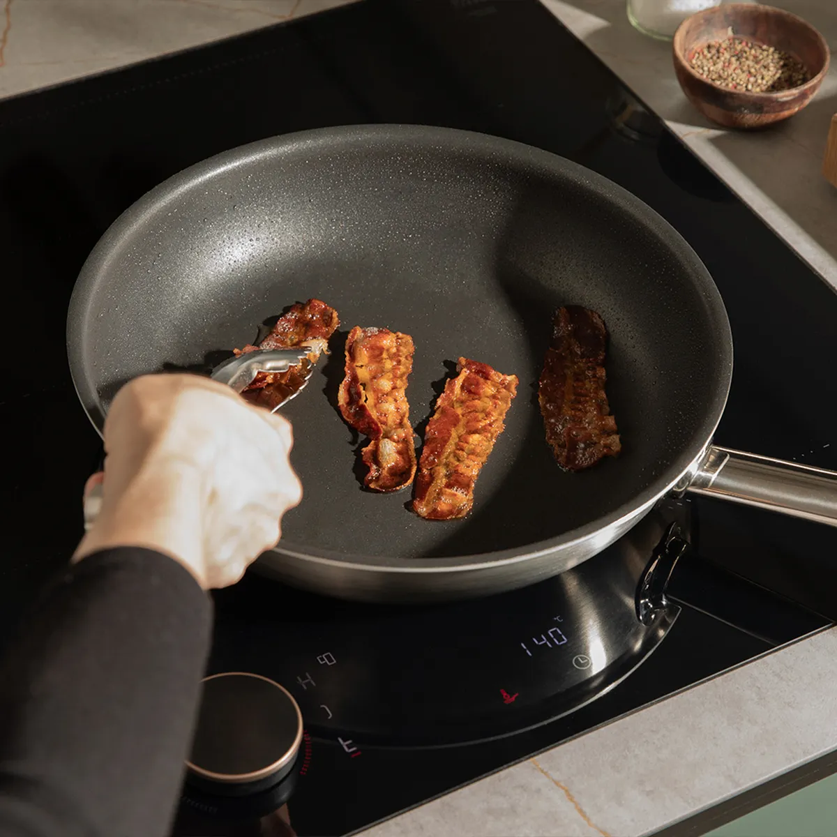 Neff induction hob with a pan of crispy bacon being turned by tongs. Digital display shows 140°C for precise cooking.