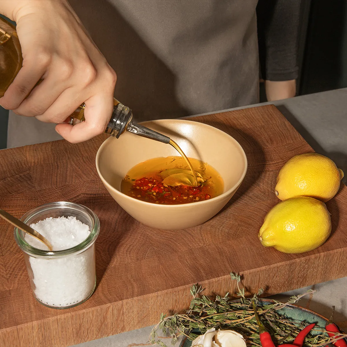 Hand pouring oil into a bowl with chili, lemons, and herbs on a wooden board. Fresh ingredients for a flavorful Neff dish.