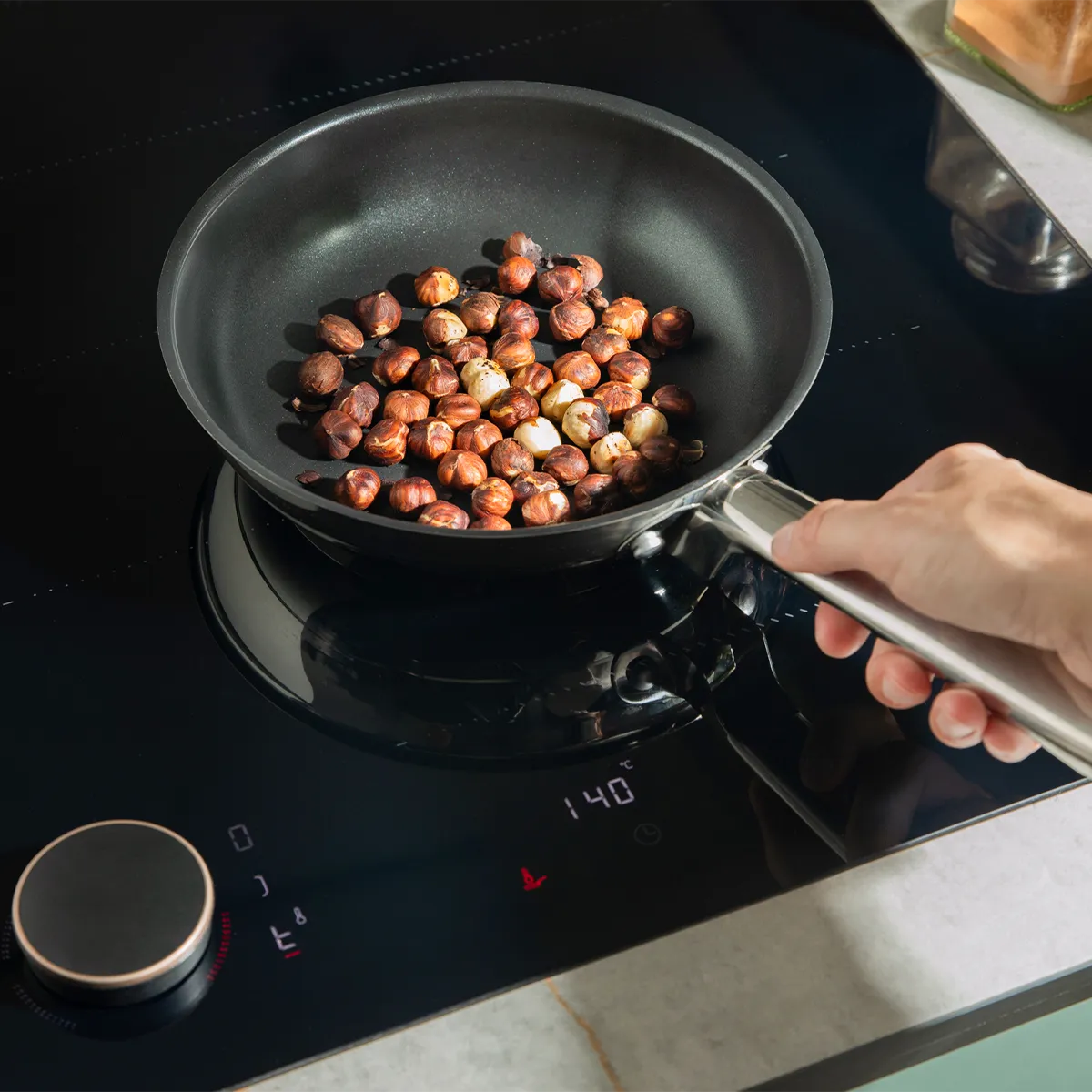 Hand roasting hazelnuts in a pan on a Neff induction hob, precisely set to 140°C. Smart cooking for perfect results.