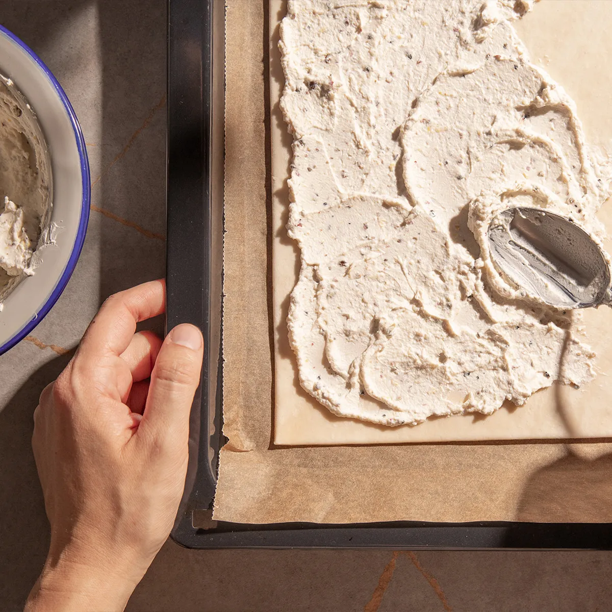Hand spreading creamy mixture on parchment paper over a baking tray with a spoon, next to a bowl of the same mixture.