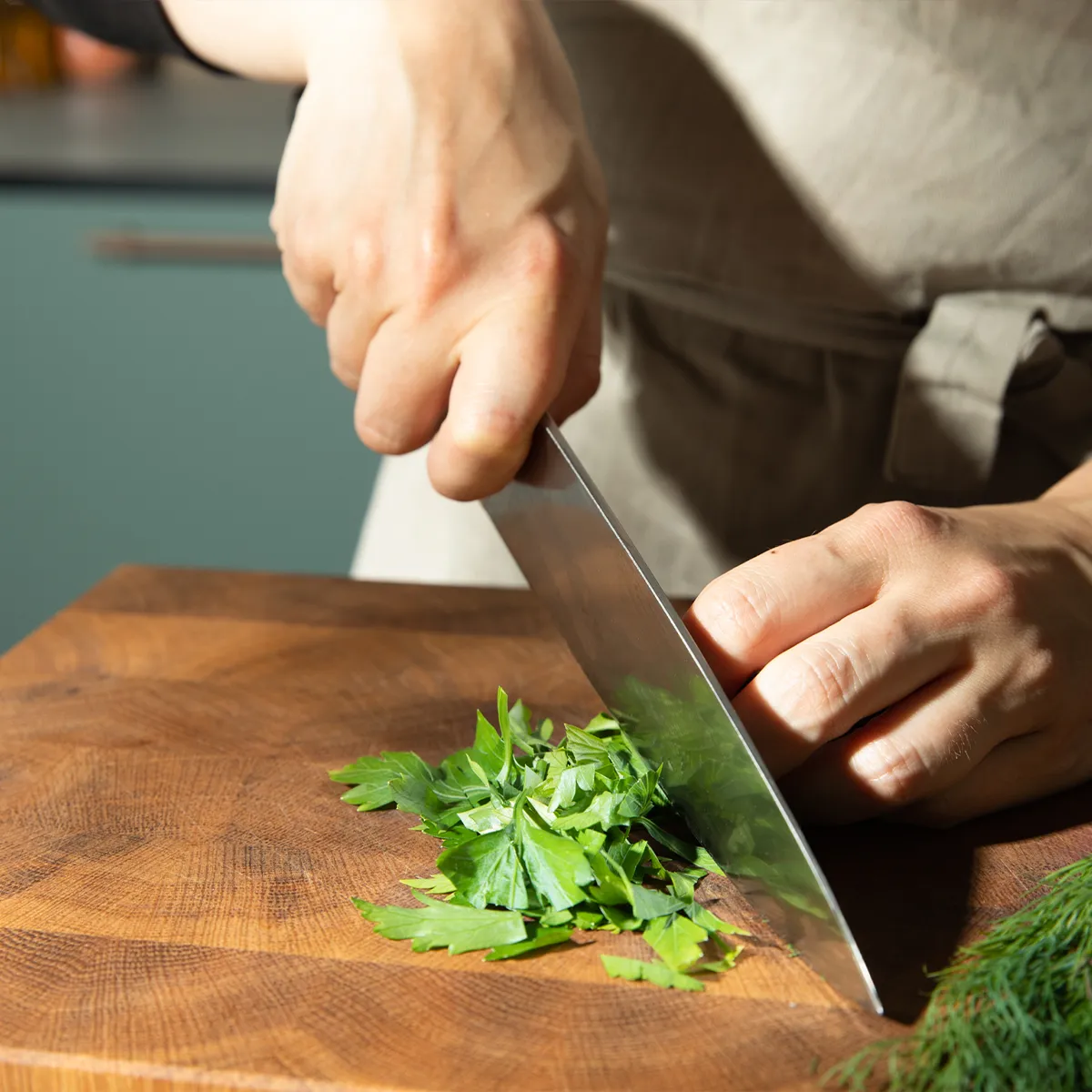 Person chopping fresh green herbs on a wooden cutting board with a large kitchen knife.
