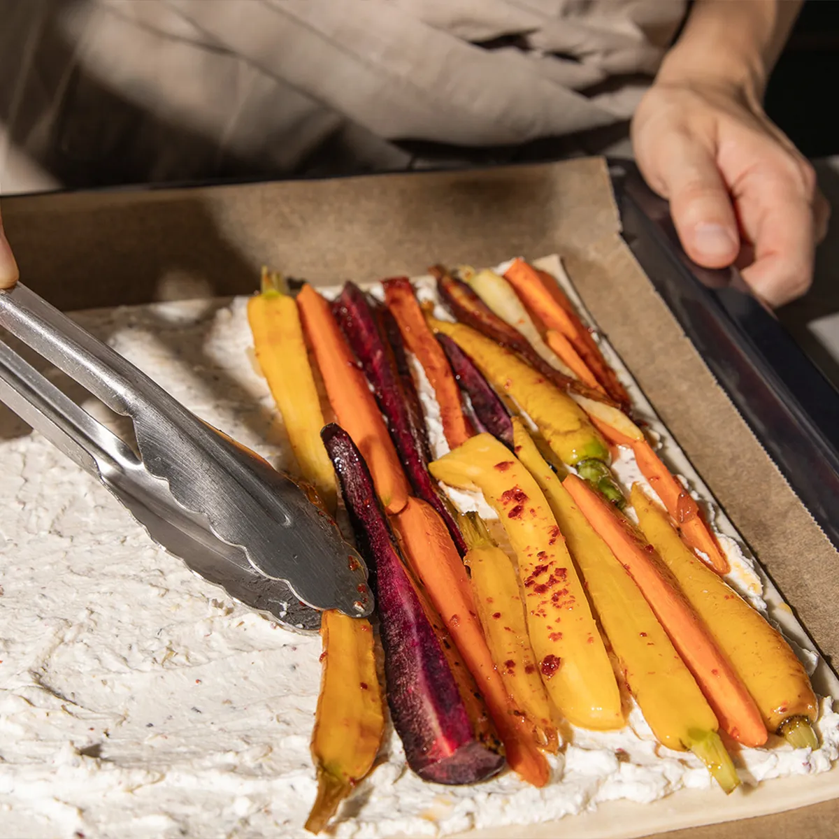 Person using tongs to arrange roasted multicolored carrots on a creamy spread over parchment-lined baking tray.