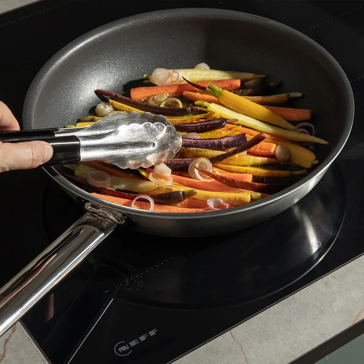 Hand using tongs to cook colorful carrot sticks and sliced onions in a frying pan on a black stovetop.