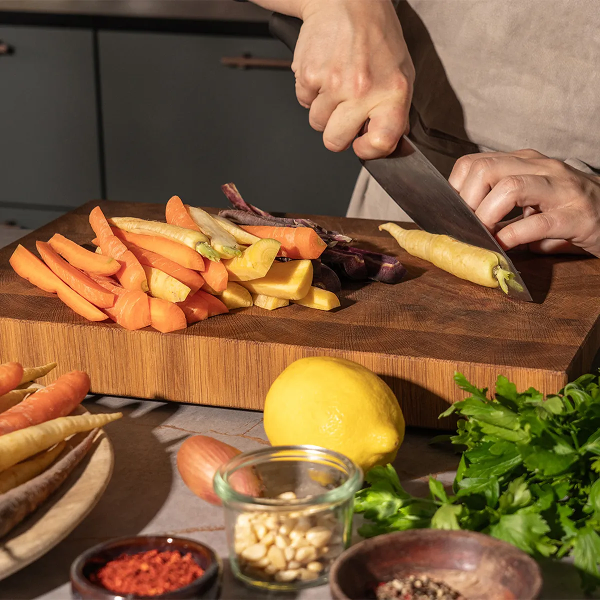 Person slicing multicolored carrots on a wooden board, surrounded by lemon, herbs, pine nuts, and spices.