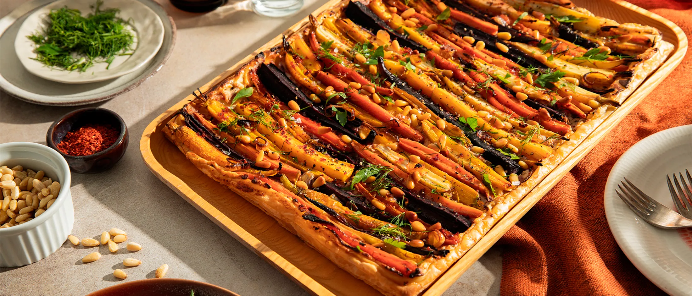 Golden puff pastry tart topped with multicolored roasted carrots, pine nuts, and fresh herbs on a wooden tray.