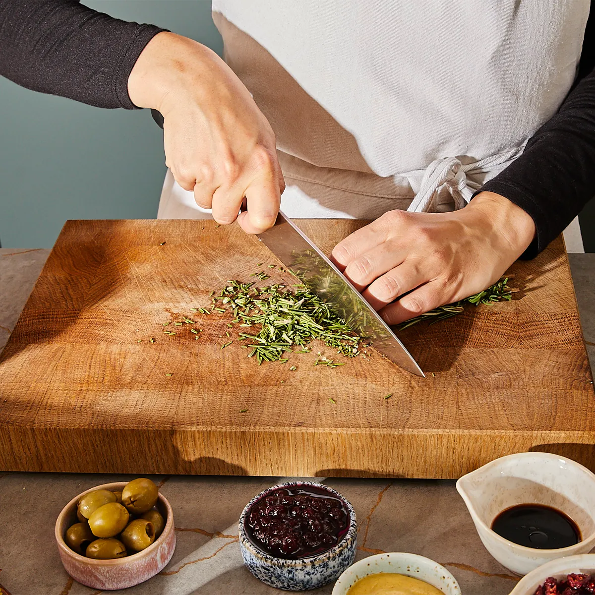 Person chopping fresh rosemary on a wooden cutting board with bowls of olives, jam, mustard, and soy sauce nearby.