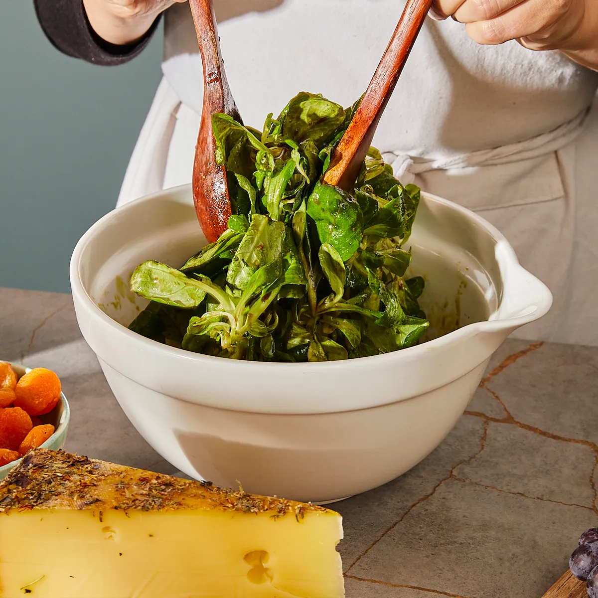 Person using wooden utensils to toss fresh greens in a white mixing bowl with cheese and carrots nearby on the counter.