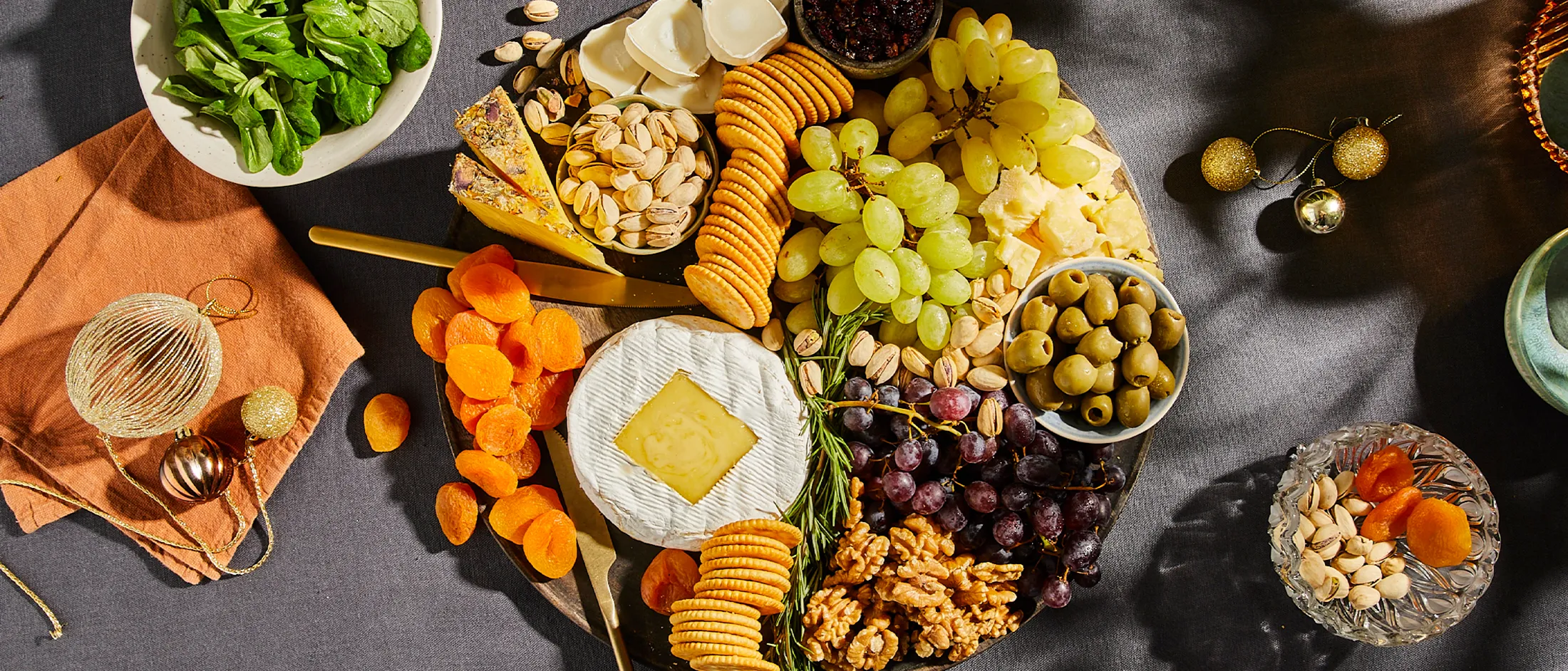 Charcuterie board with various cheeses, crackers, grapes, olives, dried apricots, pistachios, walnuts, and a bowl of spinach salad on the side.