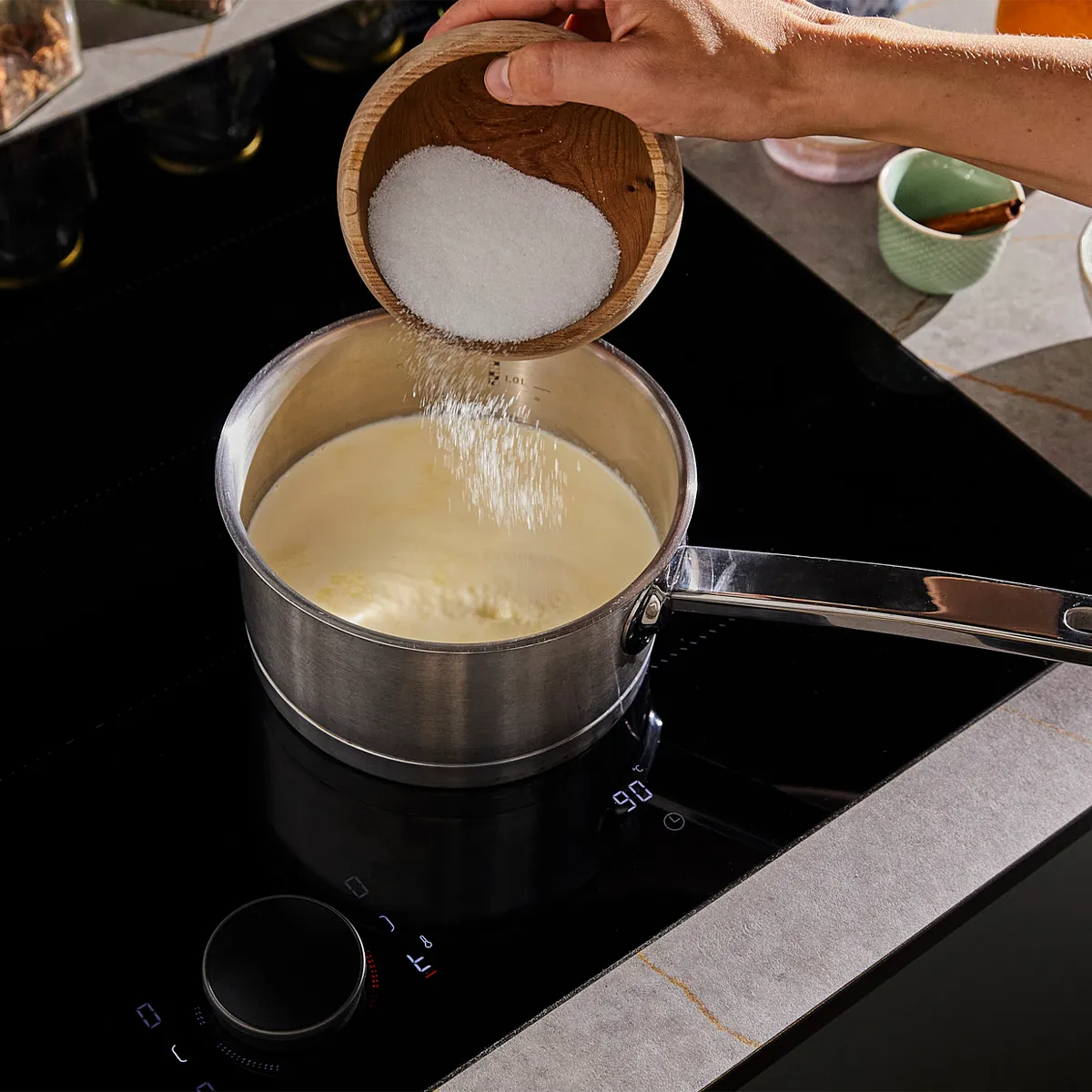 Hands pouring sugar into a pot on a stovetop, preparing a culinary dish.