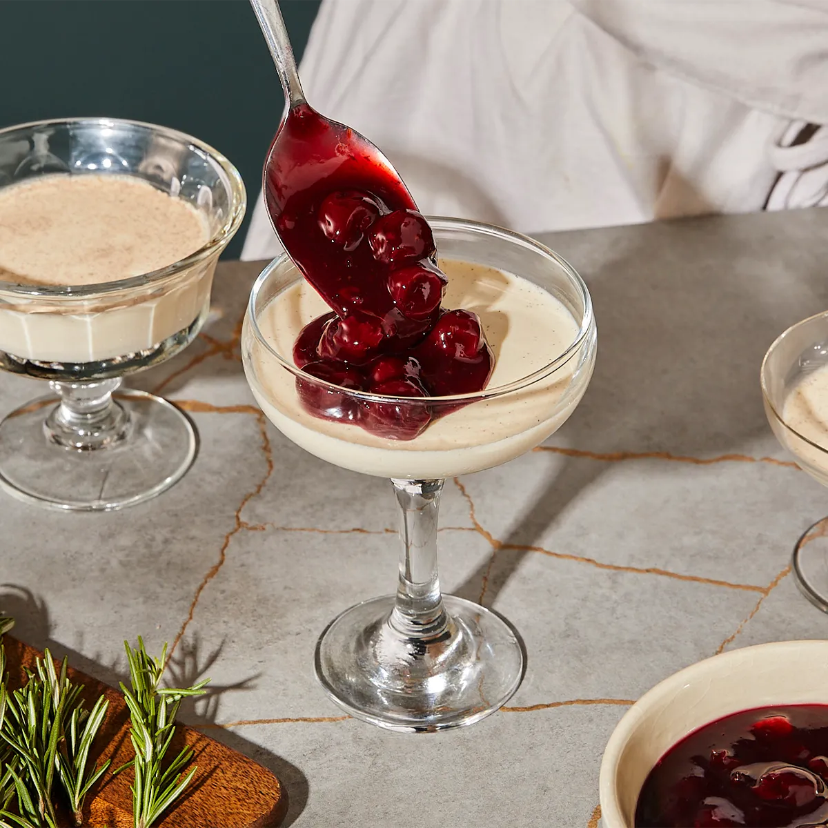 Dessert dishes with creamy custard and red berry compote, accompanied by fresh rosemary sprigs on a marble surface.