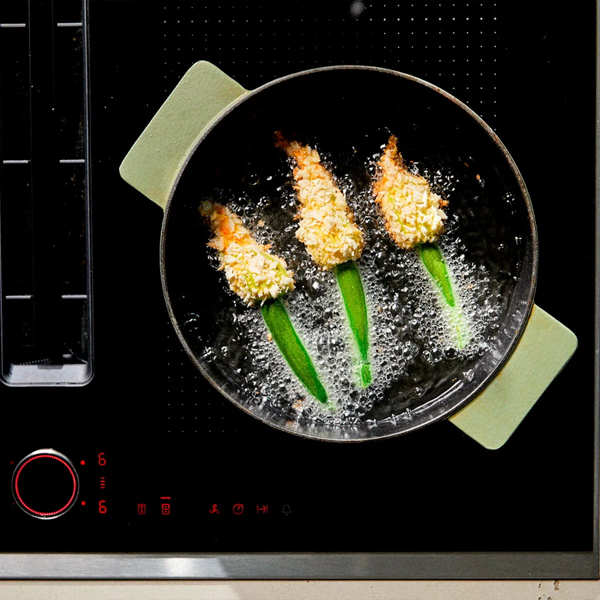 Three breaded zucchini blossoms deep-frying in a pan on a black induction hob with illuminated controls.