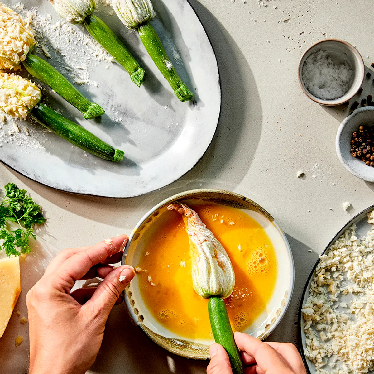 Hands coat a zucchini blossom in egg wash. Other blossoms, breadcrumbs, cheese, and spices are on the counter.