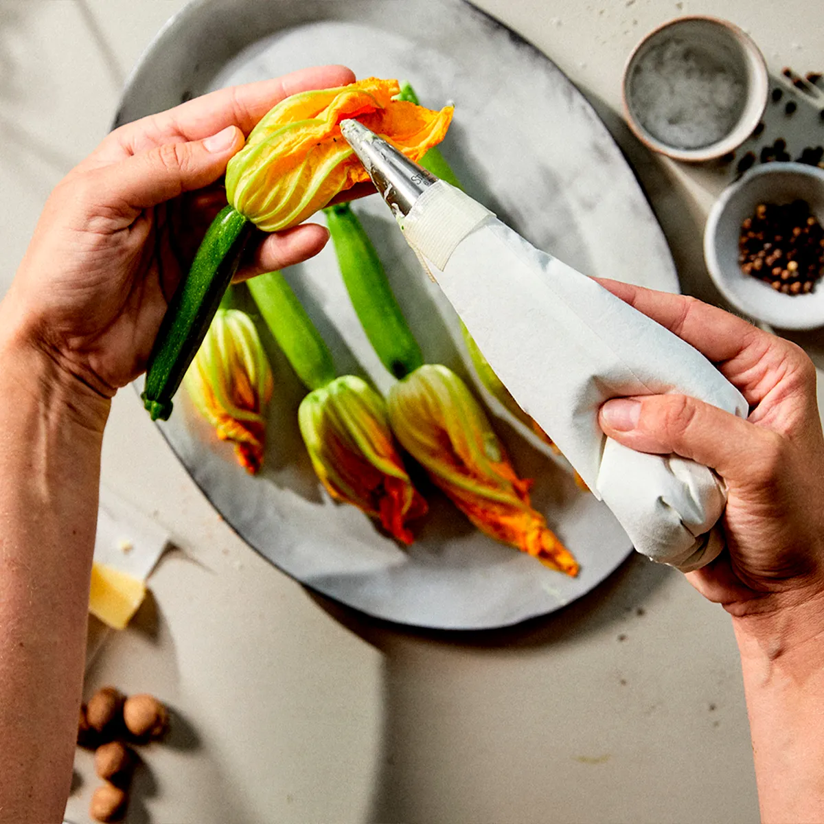 Hands pipe a filling into a vibrant zucchini blossom. Other blossoms, spices, and cheese are on the counter.