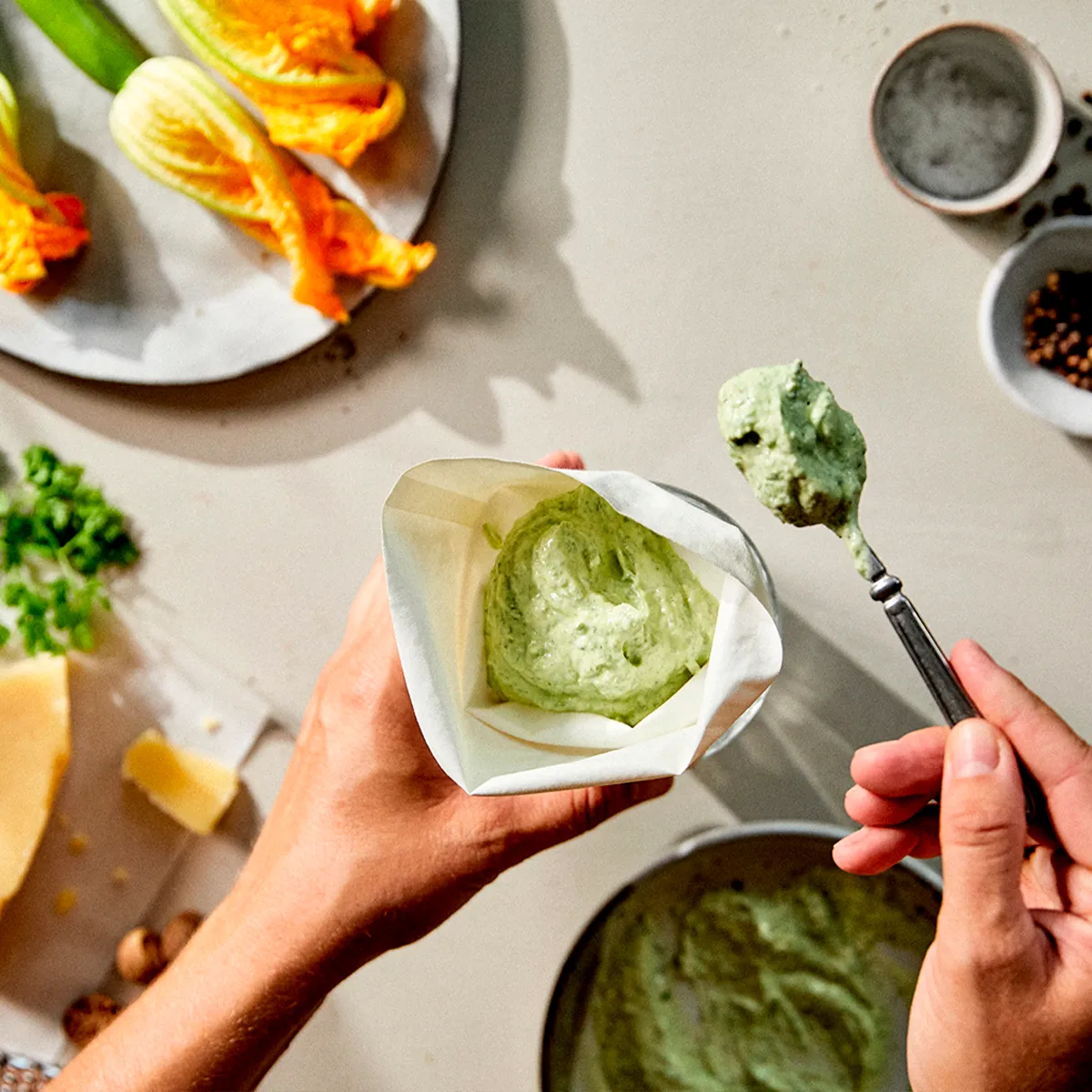 Hands fill a piping bag with green pesto, surrounded by squash blossoms, cheese, and spices on a kitchen counter.