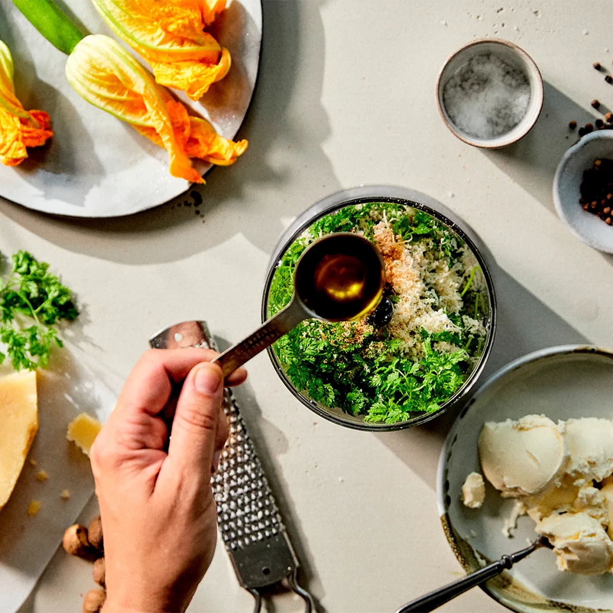  A hand adds oil to fresh herbs and grated cheese in a bowl, surrounded by squash blossoms and other cooking ingredients.