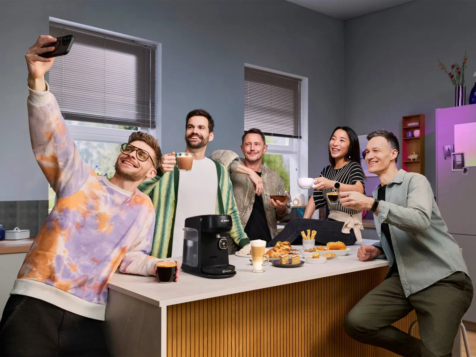 Eine vielfältige Gruppe von fünf Freund*innen in einer modernen Küche, sie lächeln und machen ein Selfie. Auf der Arbeitsplatte stehen eine Bosch TASSIMO HARMONY‑Kaffeemaschine und Gebäck; alle halten ein Getränk.