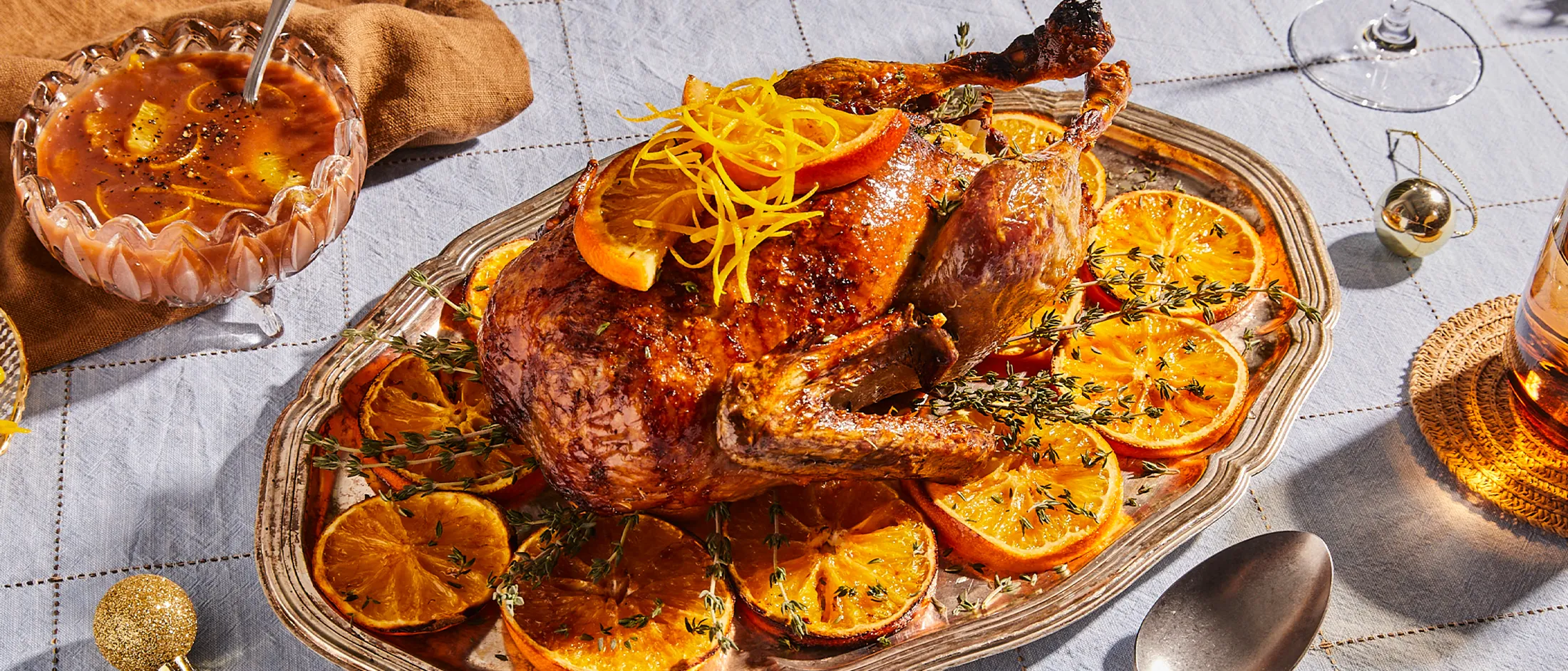 Roasted duck garnished with orange slices, served on a platter with a pie and other holiday decorations.