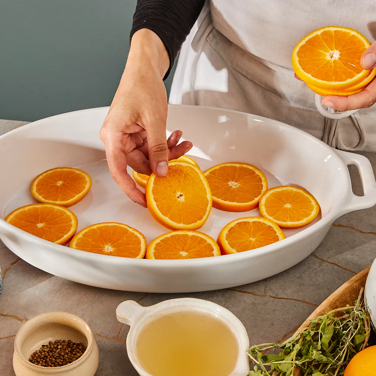Hands arranging orange slices in a baking dish, surrounded by cooking ingredients.