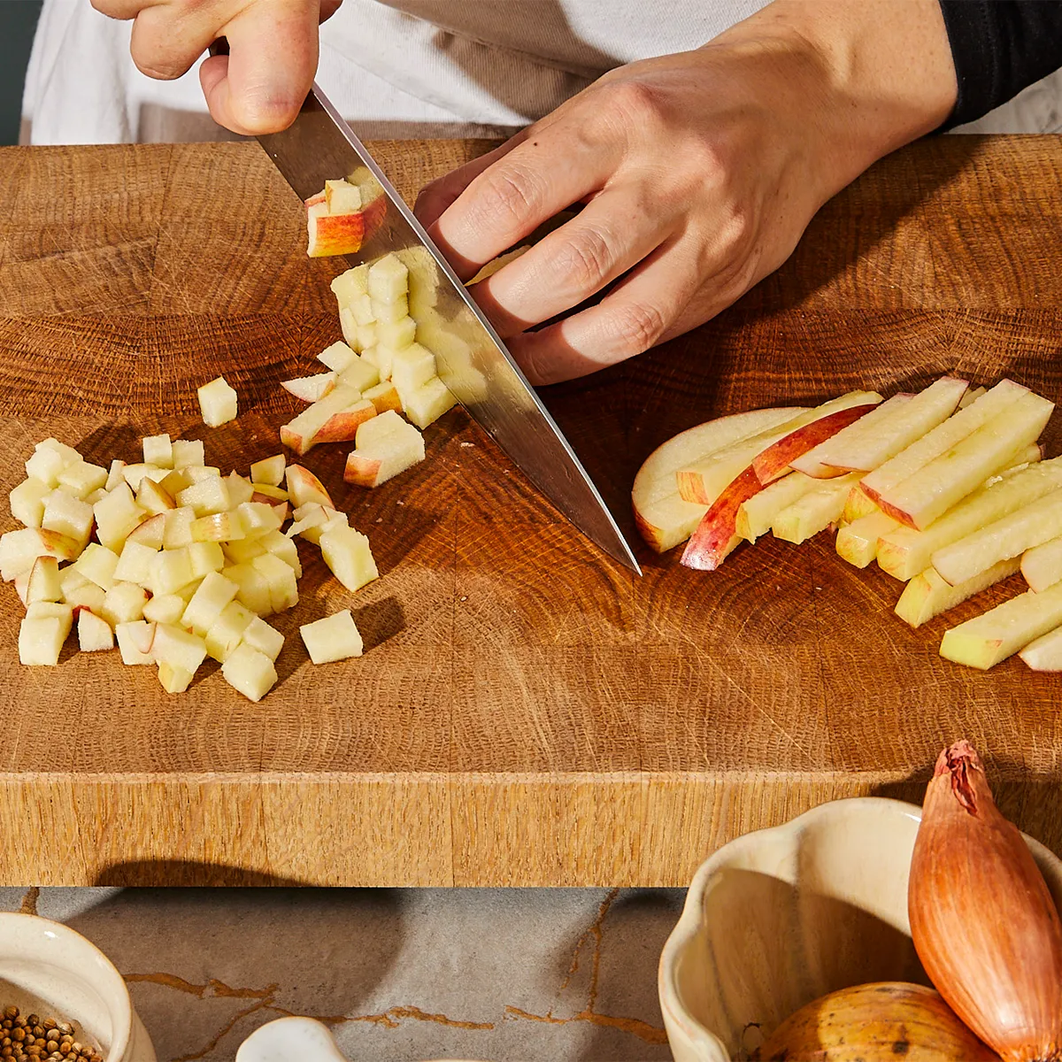 Hands chopping vegetables on a wooden cutting board, with various spices and ingredients in the background.