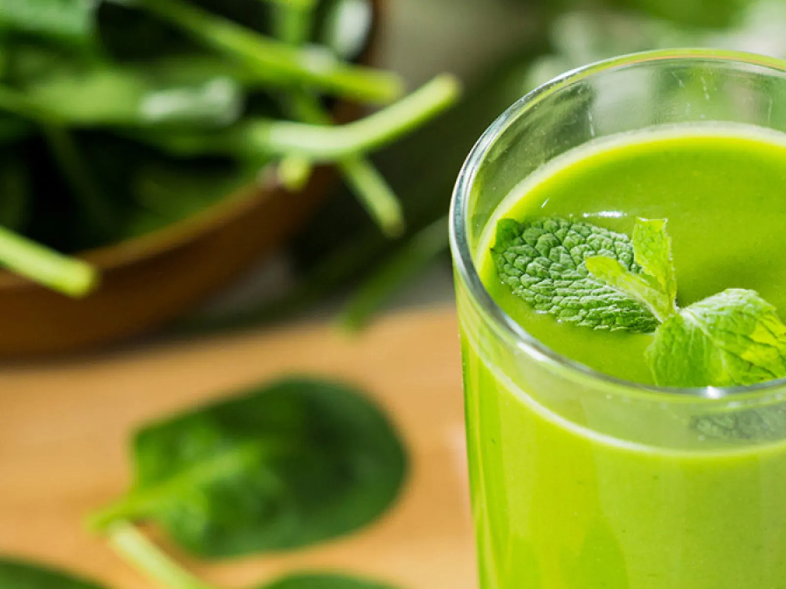 Un pahar de smoothie verde, cu o frunză de mentă deasupra și frunze de spanac așezate pe o suprafață din lemn