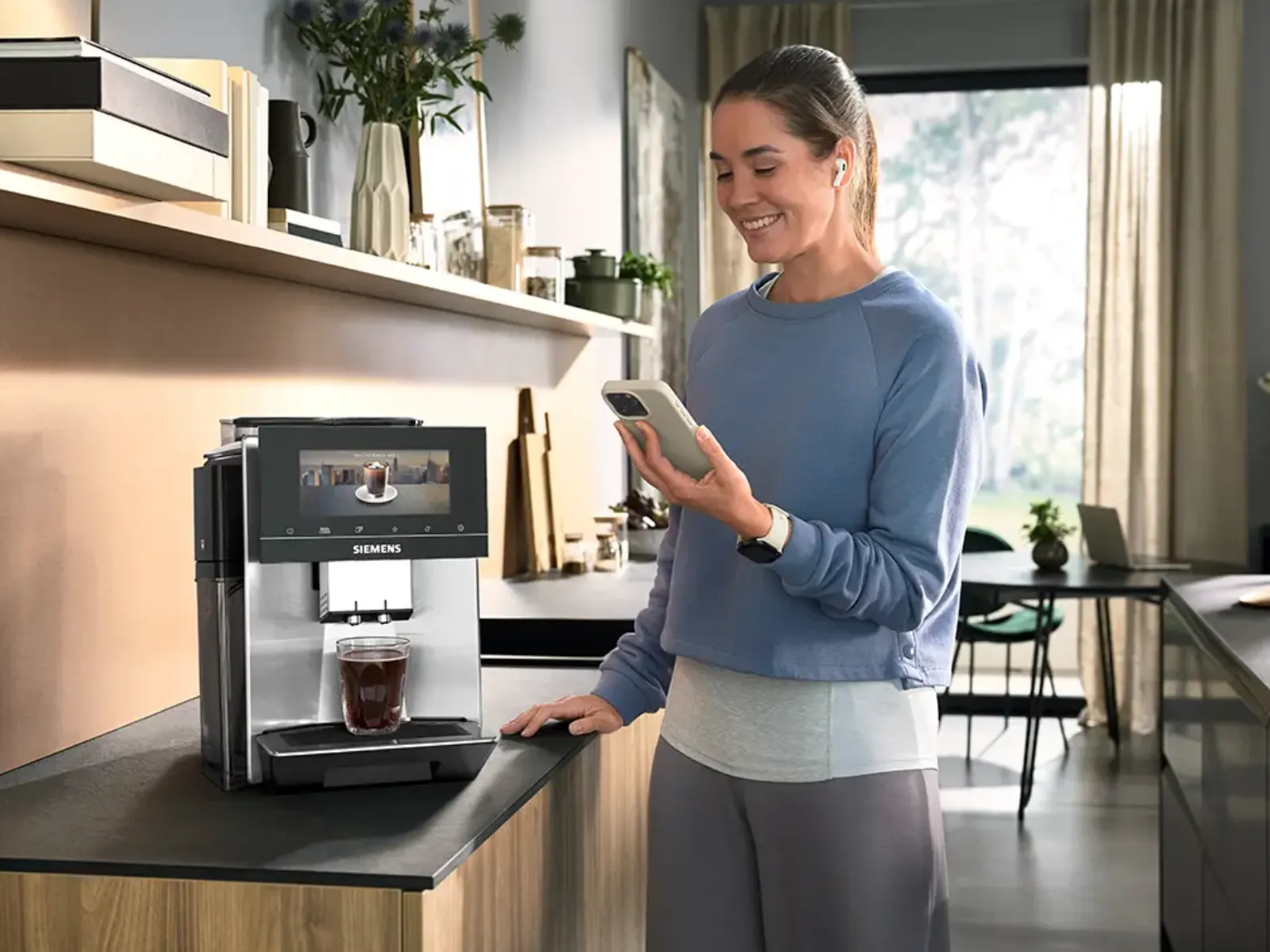 Kvinna som använder en smartphone bredvid en Siemens kaffemaskin i en modern köksmiljö.