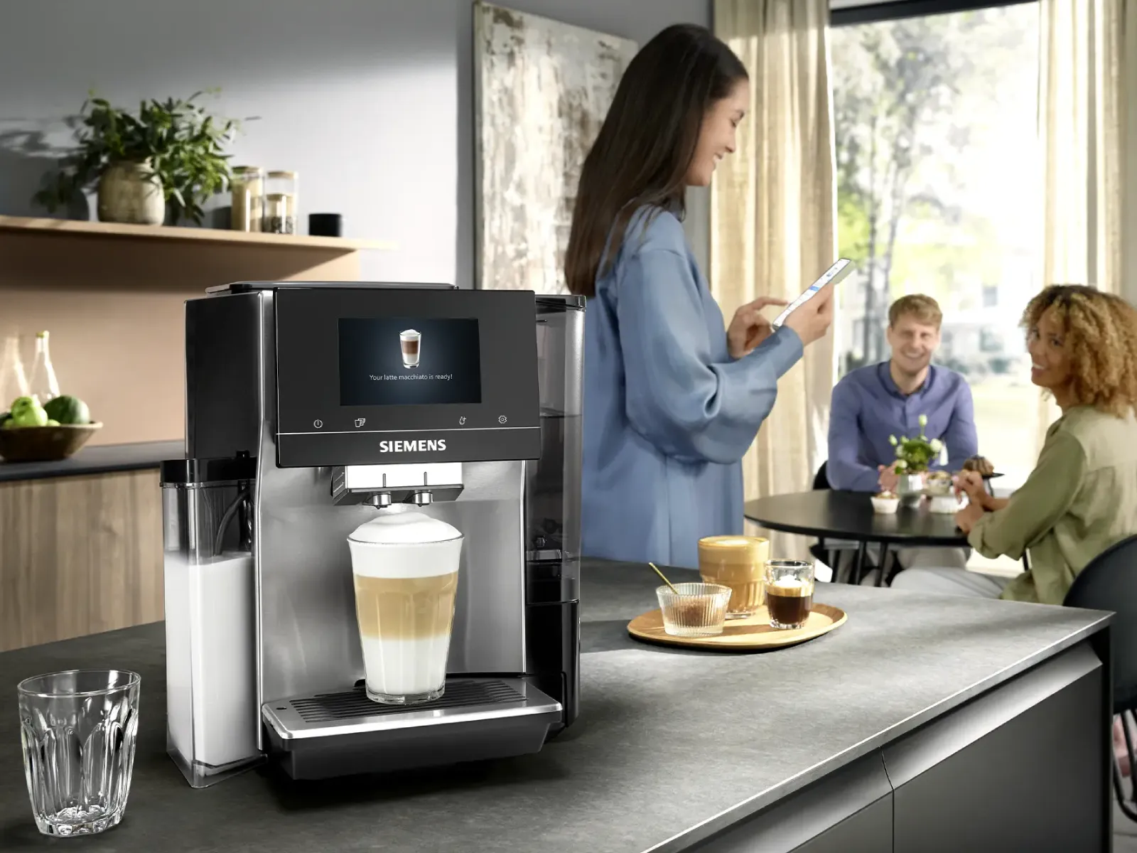 En kvinna förbereder en latte via Siemens Home Appliances-appen på sin smartphone, stående bredvid en Siemens kaffemaskin.