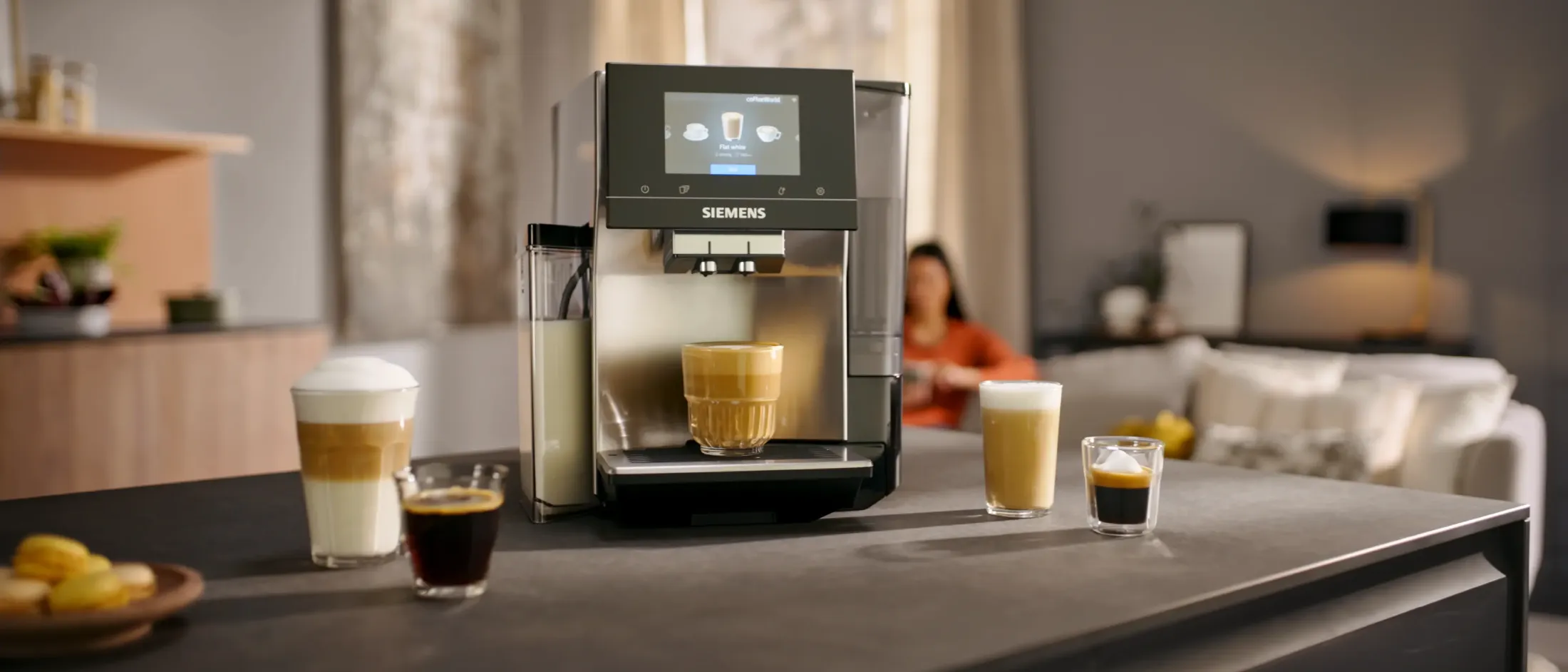 Siemens kaffemaskin på en köksö med olika kaffedrycker och macarons, en suddig person som sitter i en soffa i bakgrunden.