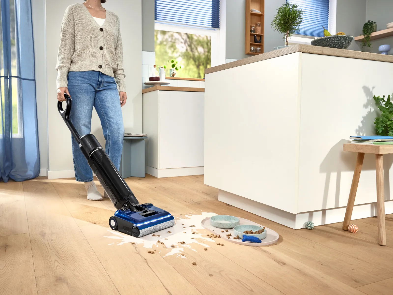 Woman cleans a spill of liquid and pet kibble on a wooden kitchen floor with a black and blue floor washer vac. Pet bowls are nearby in the modern kitchen.