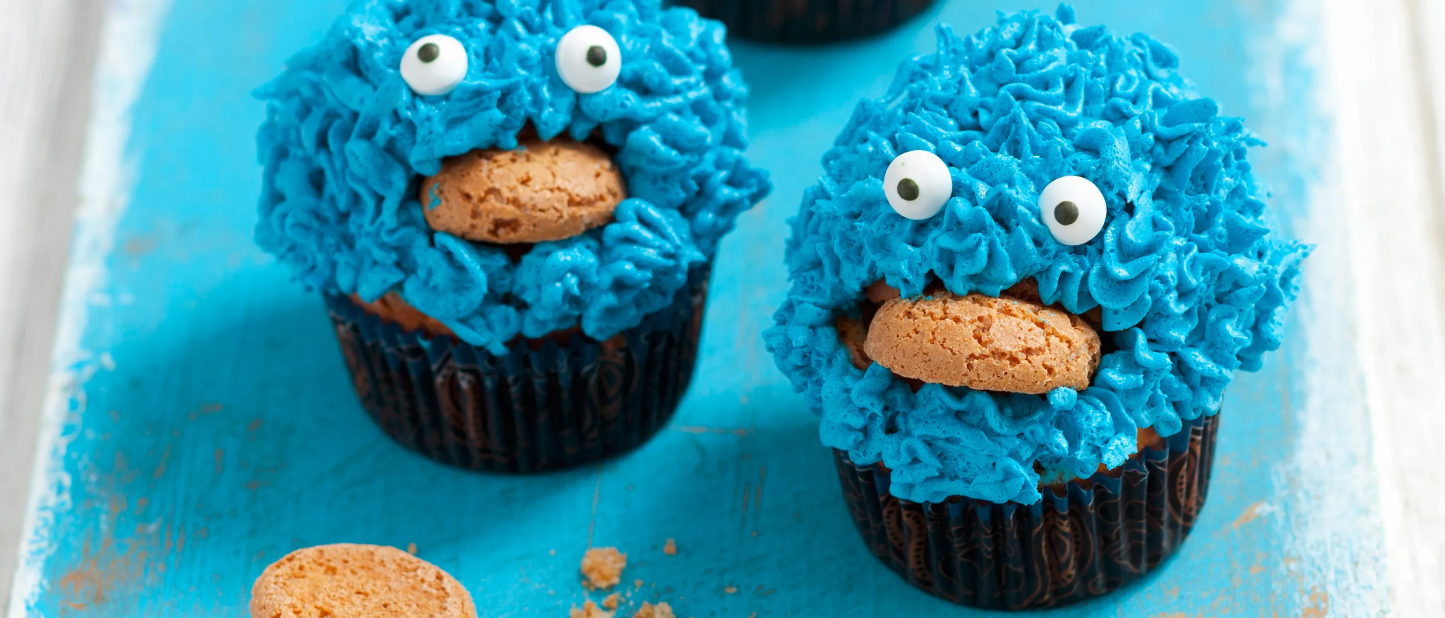 Zwei lustige Krümelmonster-Cupcakes mit blauem Frosting und einem Keks im Mund.