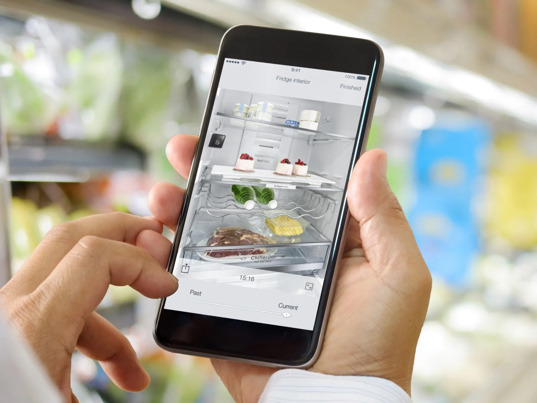 Eine Person hält ein Smartphone mit einer App für den Kühlschrank, vor dem Hintergrund einer modernen Küche und einem offenen Kühlschrank voller Lebensmittel.