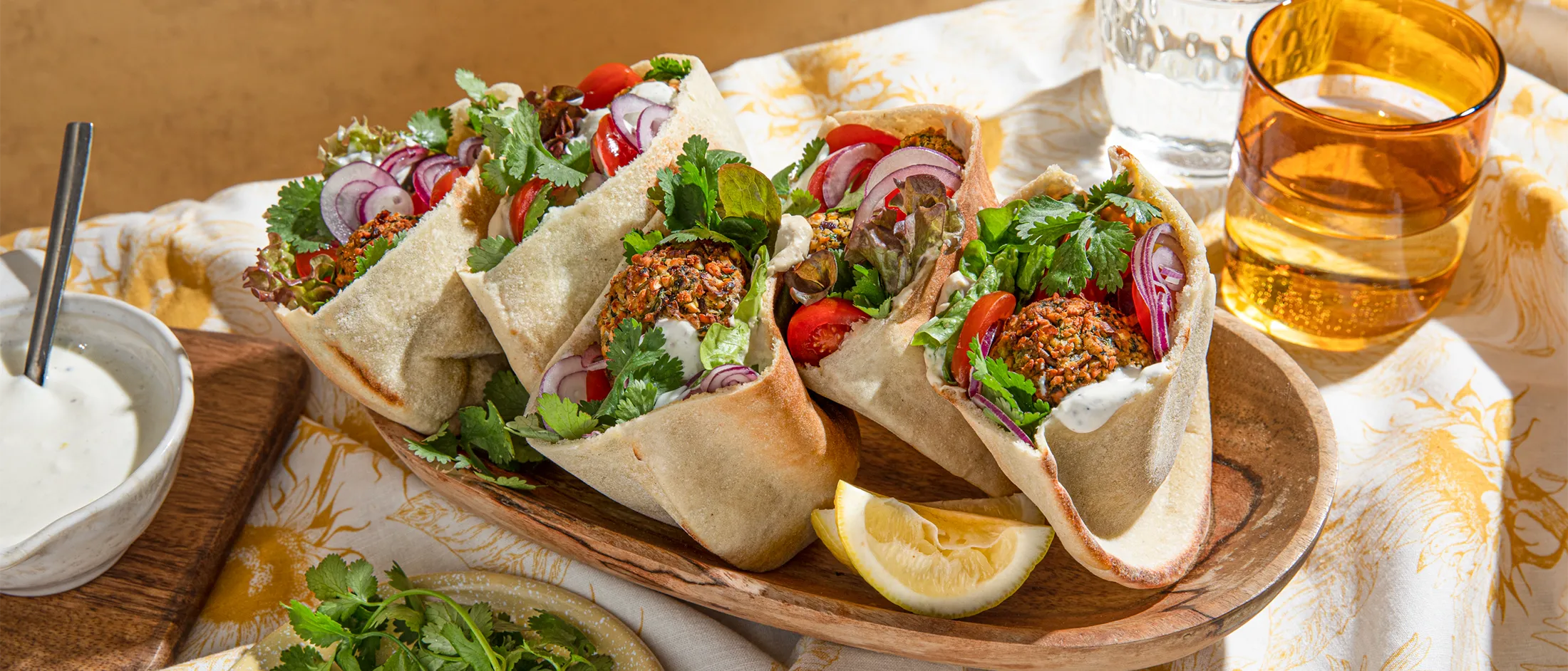 Wooden tray with four falafel pita wraps filled with lettuce, tomato, onion, herbs, and sauce, served with lemon wedges and drinks.