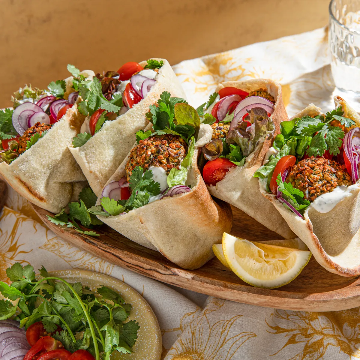 Wooden tray with four falafel pita wraps filled with lettuce, tomato, onion, herbs, and sauce, served with lemon wedges and drinks.