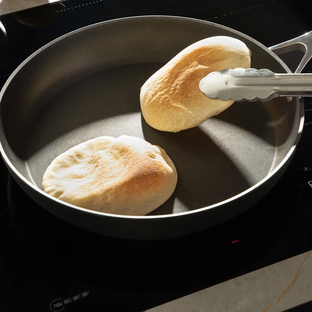 Two pieces of pita bread toasting in a nonstick pan on a stovetop, one held with metal tongs.