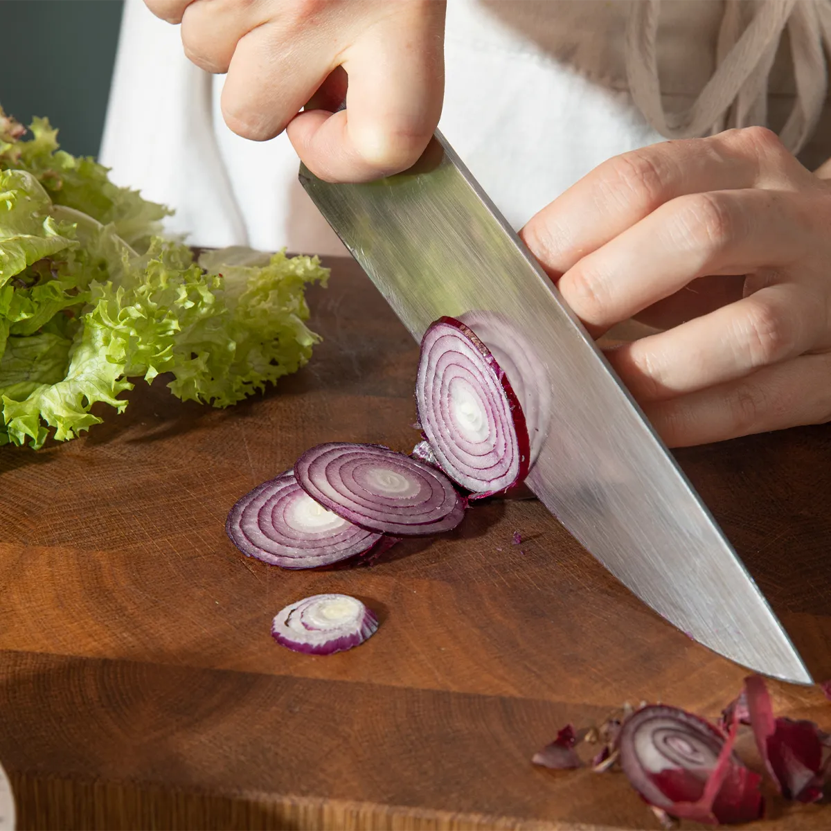 Hands slicing red onion on a wooden cutting board with green lettuce nearby.
