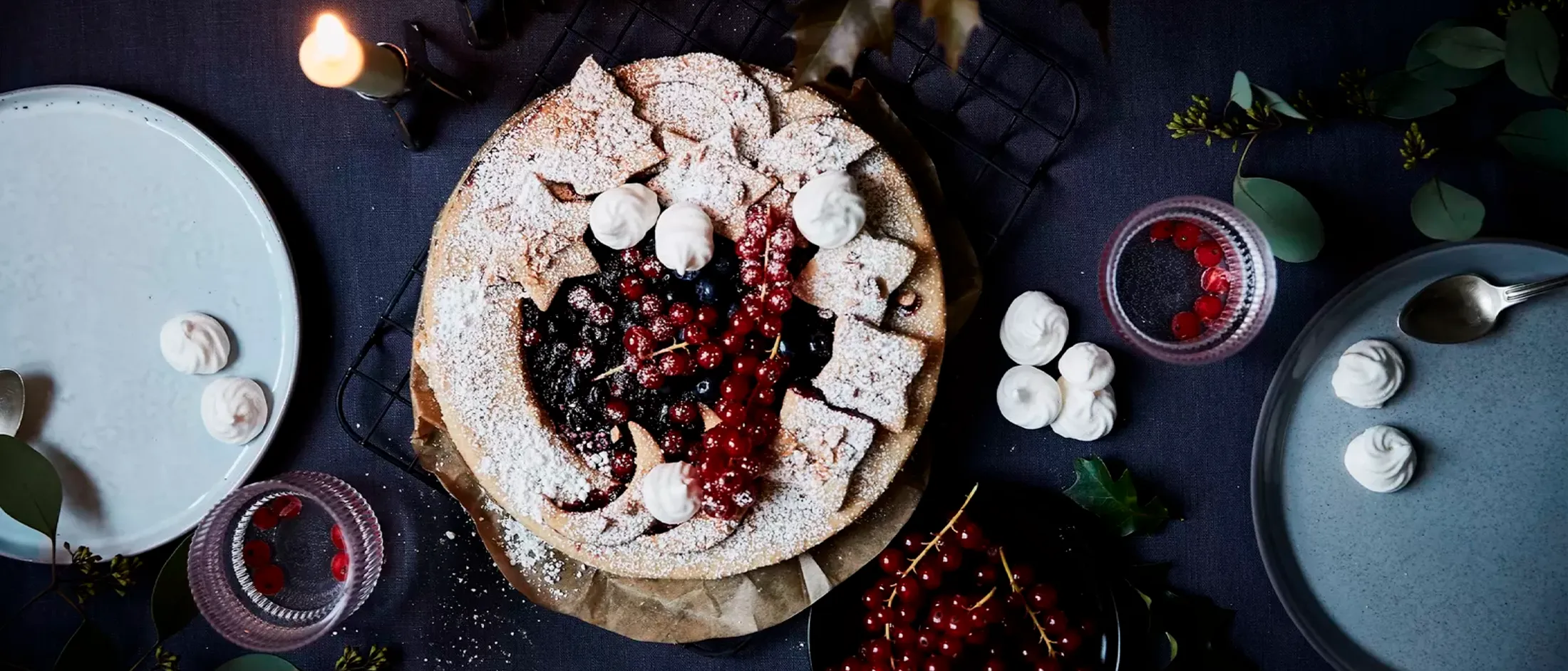 Top-down view of a berry tart dusted with powdered sugar, surrounded by plates with meringues, red currants in glasses, and a lit candle.