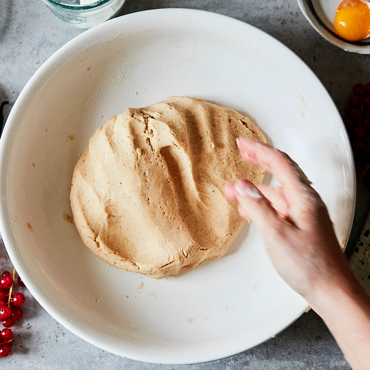 Hand pressing into dough in a white bowl on a kitchen counter with an egg yolk and red currants nearby.
