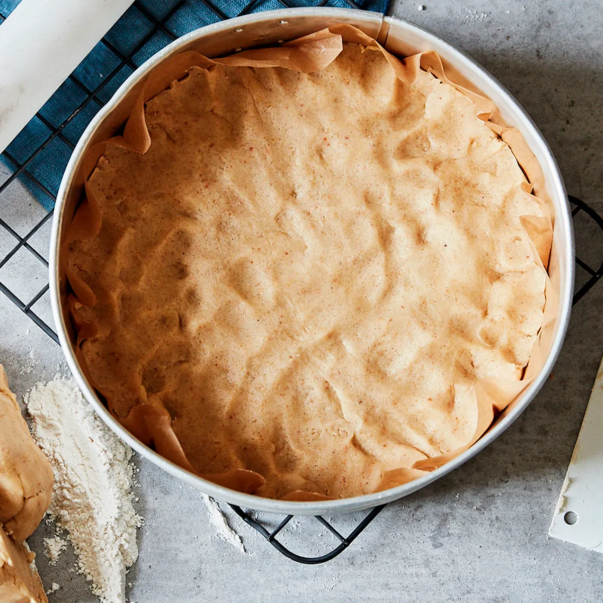 A raw pie crust in a parchment-lined round baking pan on a cooling rack. Flour and extra dough are on the counter.