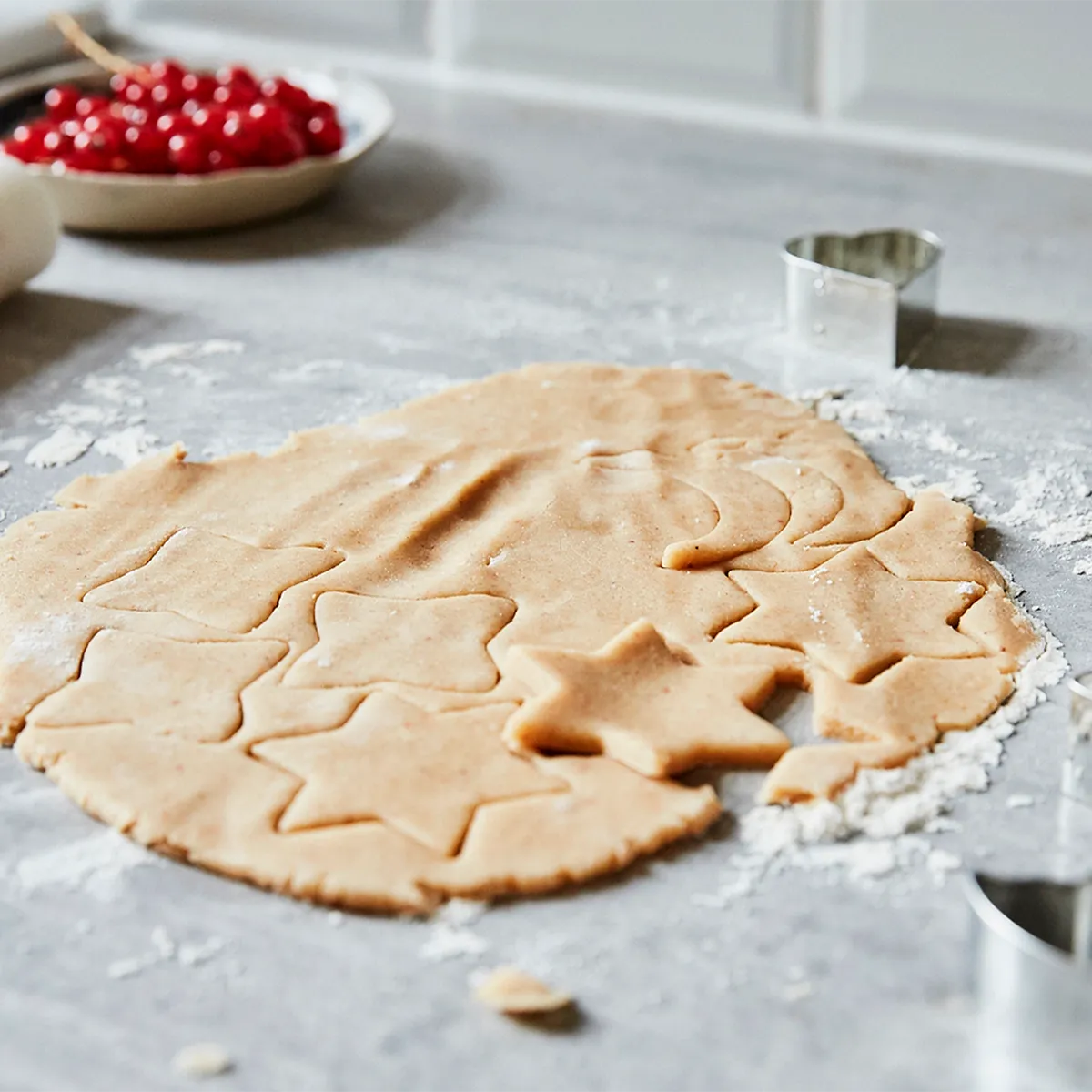 Cookie dough with star and moon cutouts on a floured counter, with cookie cutters and red currants in the background.