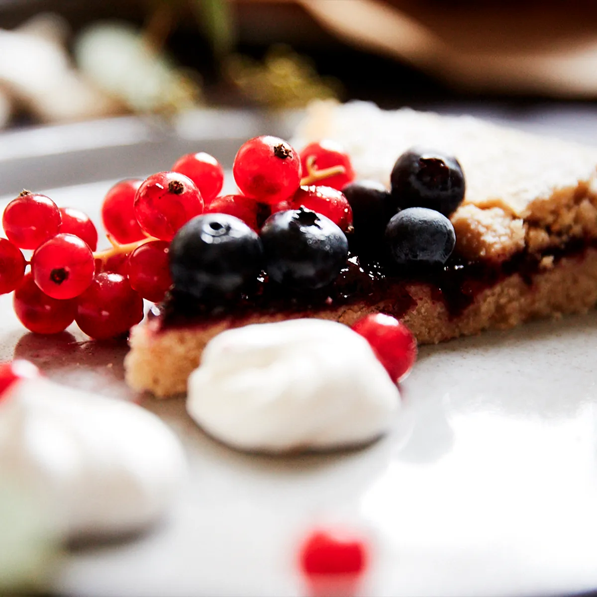 A slice of berry pie topped with fresh red currants, blueberries, and a dollop of cream on a white plate.