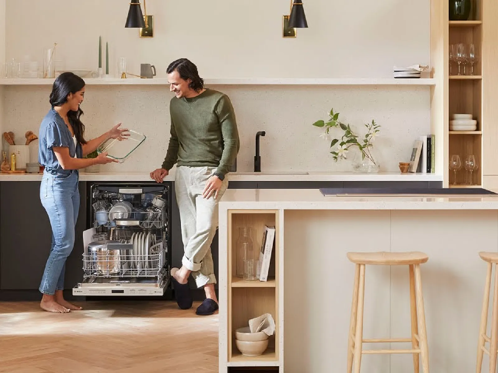 Couple unloading a dishwasher in a modern kitchen with wooden stools, plants, and open shelves.