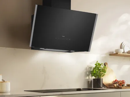 Moderne schwarze, angewinkelte Küchen-Dunstabzugshaube über einem eleganten Kochfeld mit Kräutern und Küchenutensilien auf der Arbeitsplatte und im Regal.
