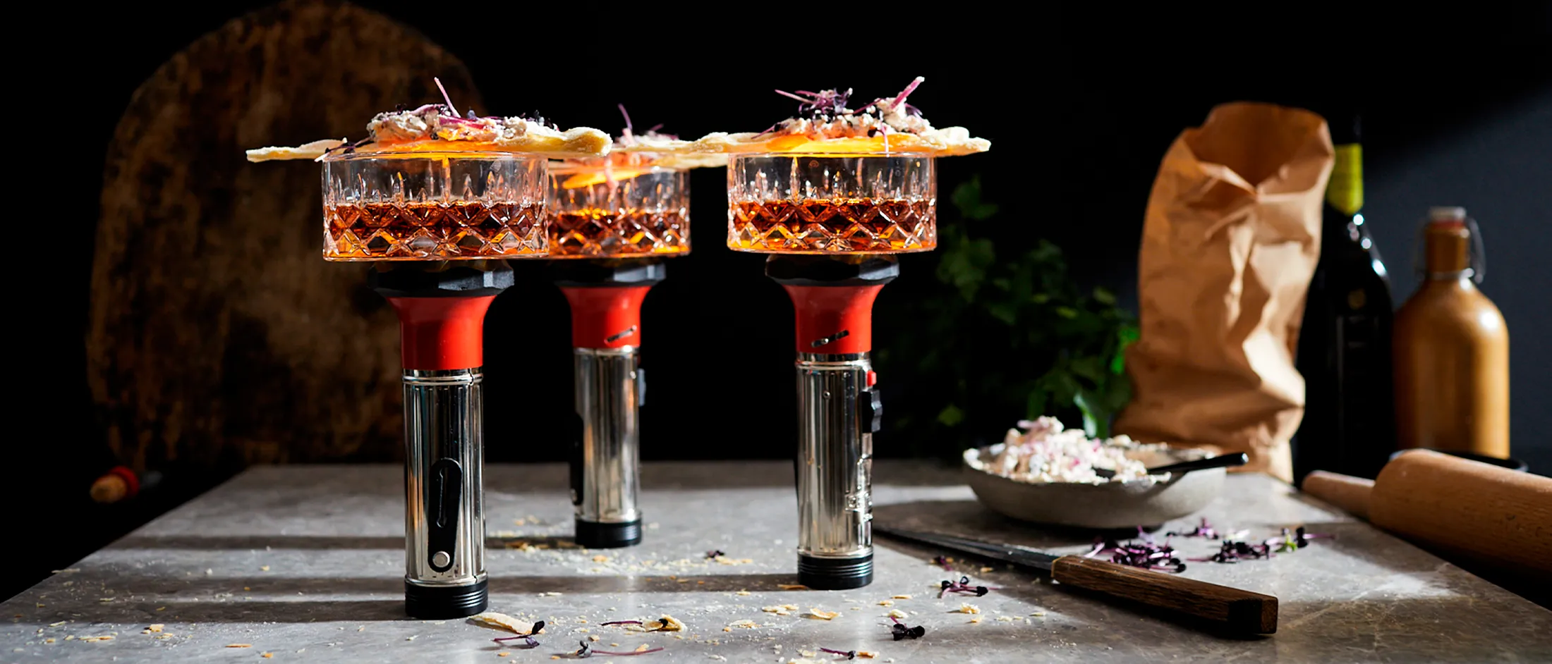 Cocktail glasses filled with amber liquid, garnished with citrus slices, on a dark background with various bar tools.