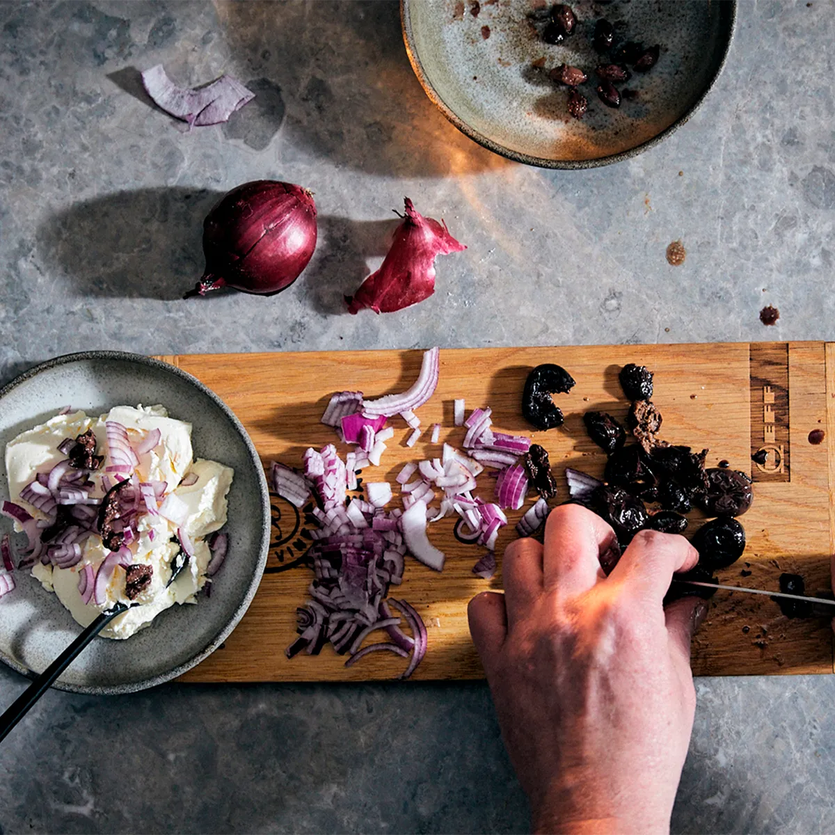 Hands chopping vegetables on wooden cutting board, surrounded by various ingredients on a gray surface.