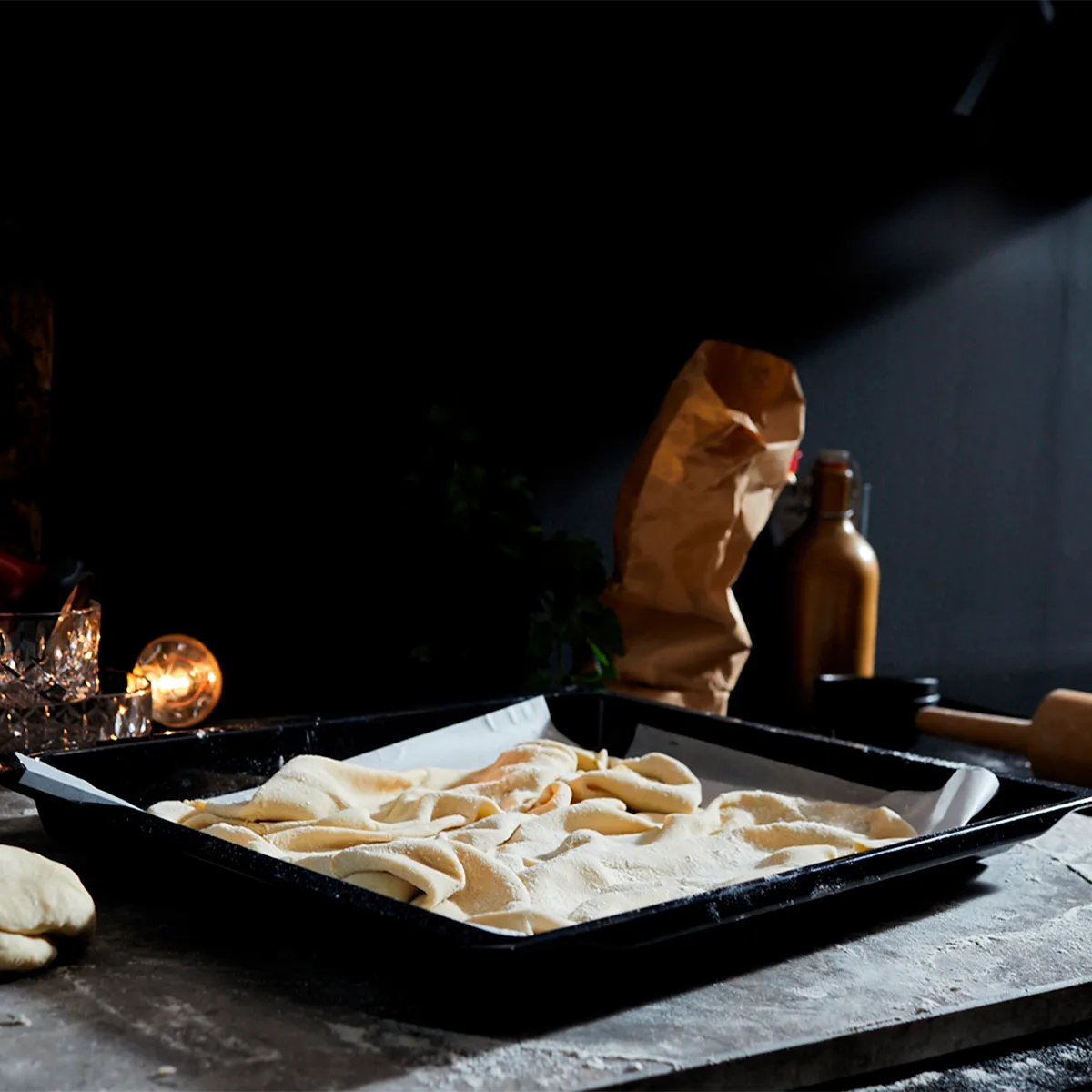 Dough, rolling pin, baking tray, spices, and cooking utensils on a dark kitchen counter.