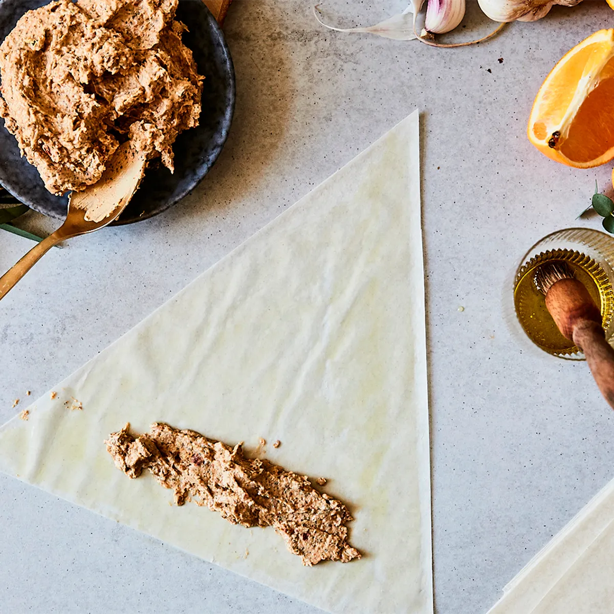Triangle of pastry dough with a nut mixture spread on it, a bowl of the mixture, garlic, orange slice, and a honey brush nearby on a countertop.
