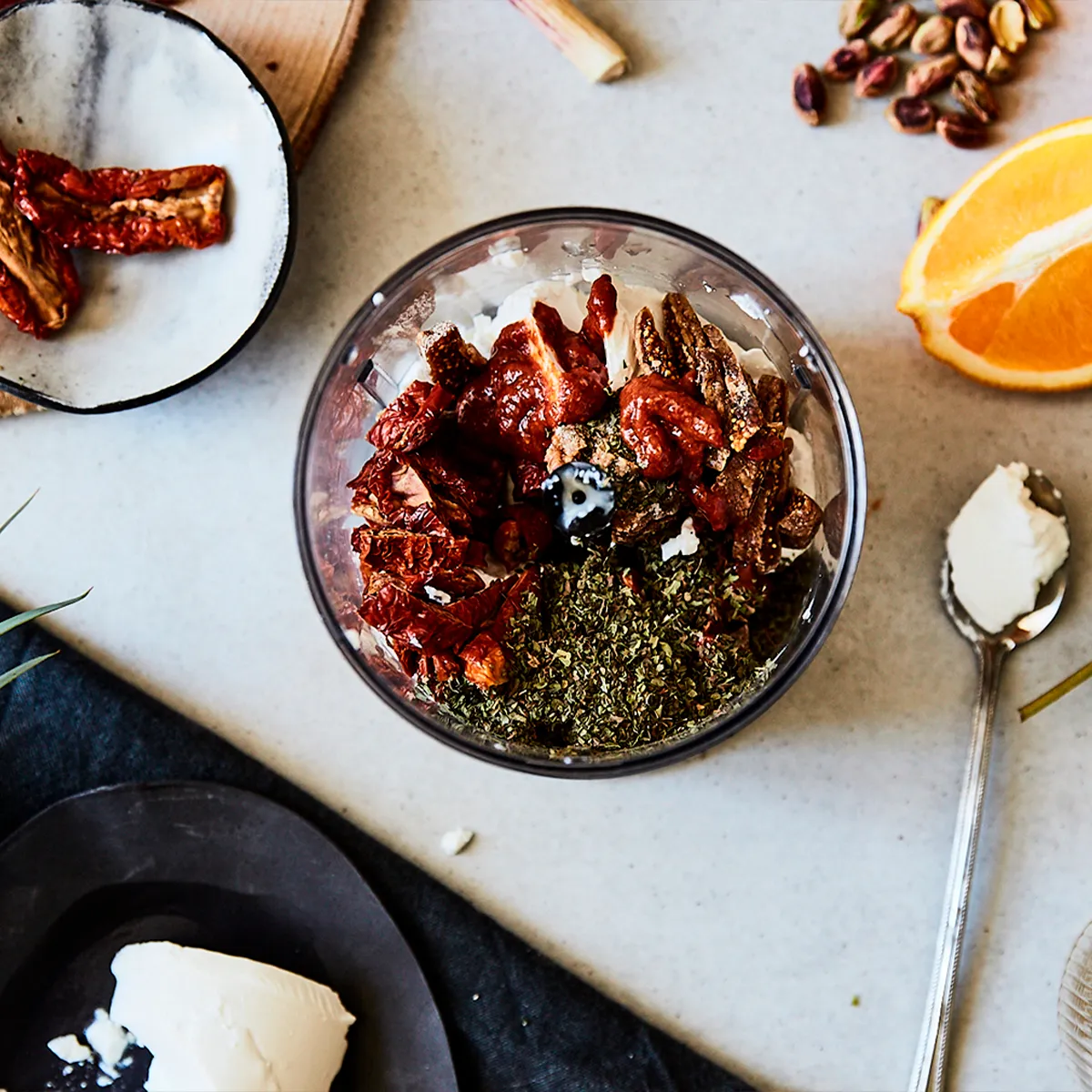 Top view of dried tomatoes, herbs, cream cheese, and orange slices arranged for food preparation on a kitchen countertop.