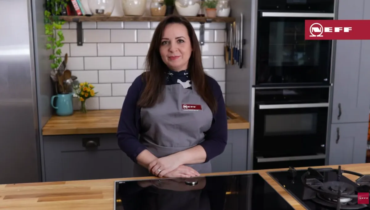 Woman with long dark hair in a kitchen, wearing a gray apron and black shirt, standing behind a countertop with a stovetop in front.