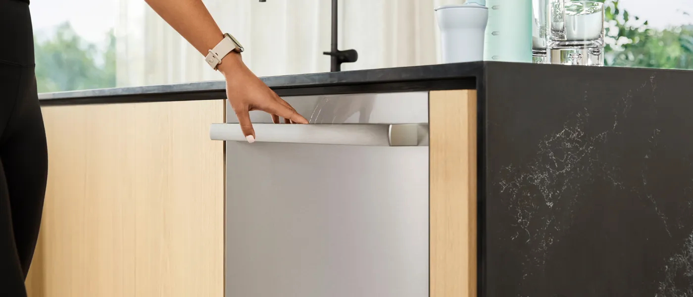 A person’s hand pulls open a built-in stainless steel dishwasher in a modern kitchen with light wood and dark marble counters.