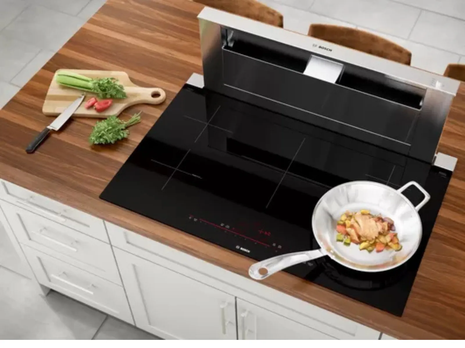 Modern kitchen with a wooden countertop, built-in black induction cooktop, downdraft vent, and a frying pan cooking vegetables.