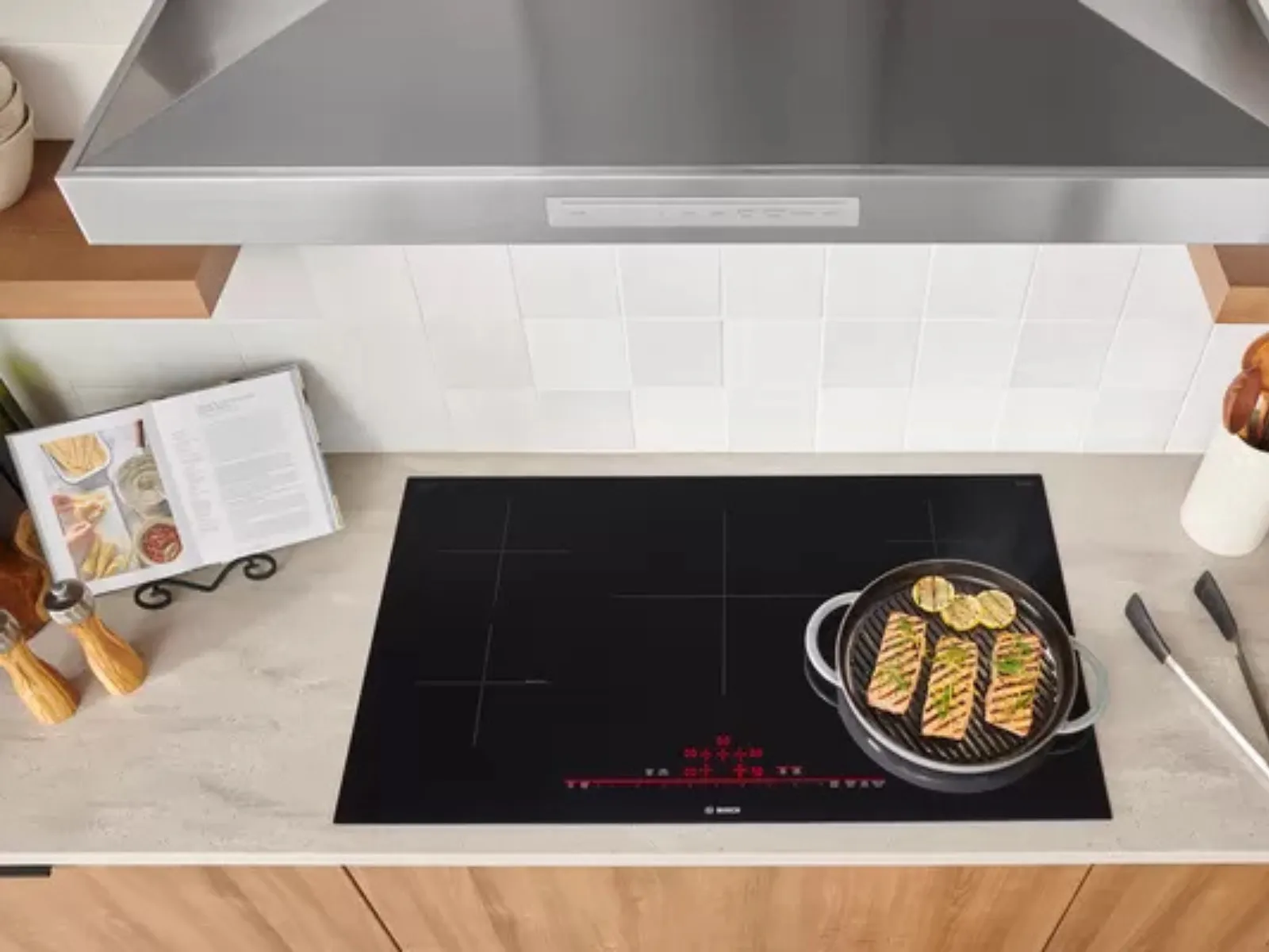 Top-down view of a modern kitchen countertop featuring an induction cooktop with fish and lemon slices cooking on a pan.